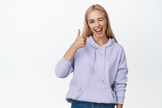 Happy blond girl winking and smiling showing thumbs up like and approve complimenting you standing over white background