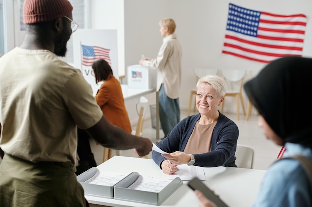 Happy blond mature woman passing ballot paper to African American man