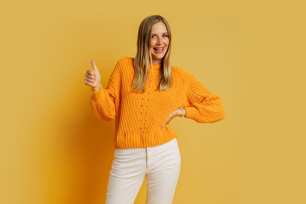 Фото Счастливая белокурая женщина в оранжевом стильном осеннем свитере, позирующем на желтом. показываю знак ок.