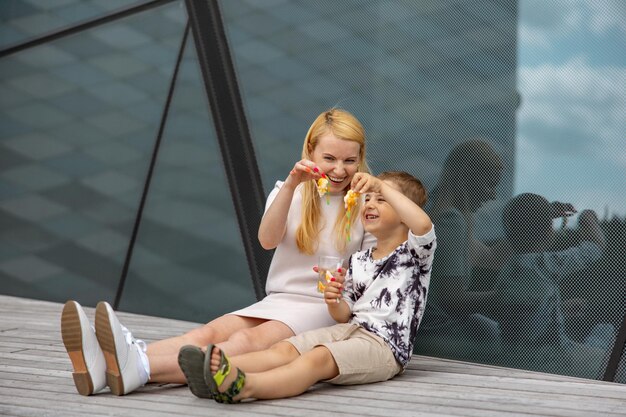 Happy blond woman and little boy sitting on terrace and eating sweets Mother and son enjoy time together Positive young mom playing spending time with her cute child laughing having fun Family