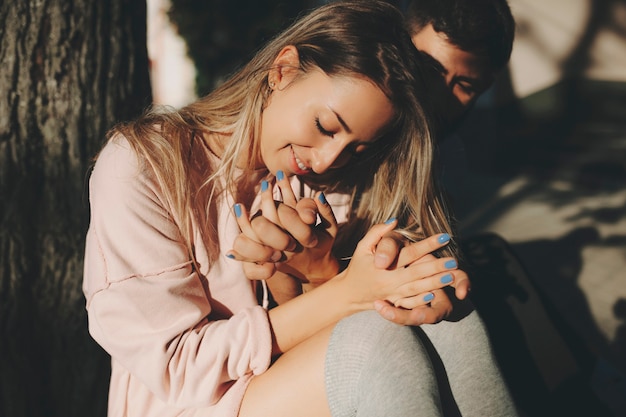 Happy blond woman with man sitting near tree in sunlight cuddling with love