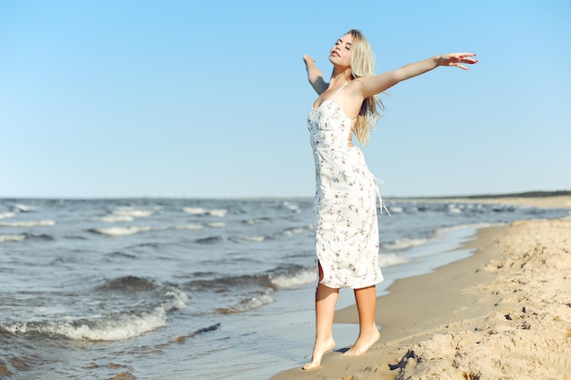 Happy blonde beautiful woman on the ocean beach standing in a white summer dress, open arms.