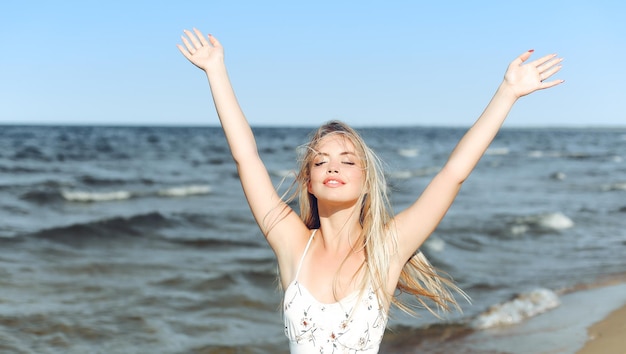 Happy blonde beautiful woman on the ocean beach standing in a white summer dress, raising hands.