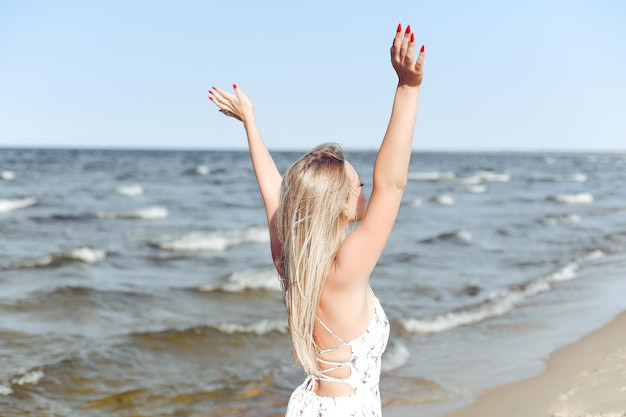 Happy blonde beautiful woman on the ocean beach standing in a white summer dress, raising hands.