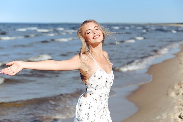Счастливая блондинка красивая женщина на берегу океана стоит в белом летнем платье и поднимает руки.