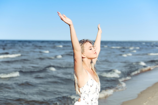 Happy blonde beautiful woman on the ocean beach standing in a white summer dress, raising hands