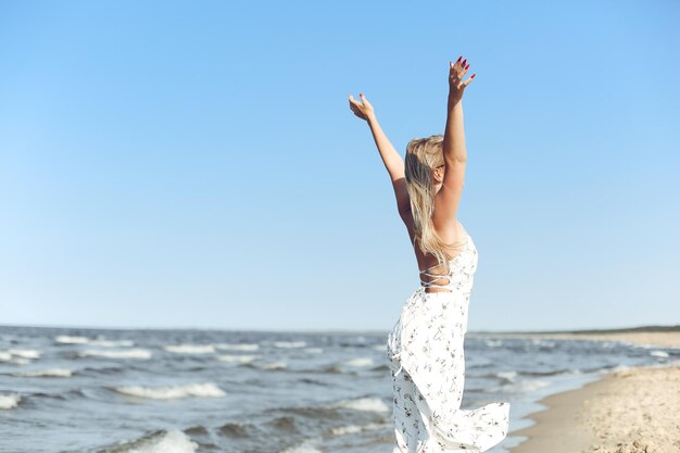 海のビーチで白い夏のドレスとサングラスを着て立ち、手を上げる幸せな金髪の美しい女性。