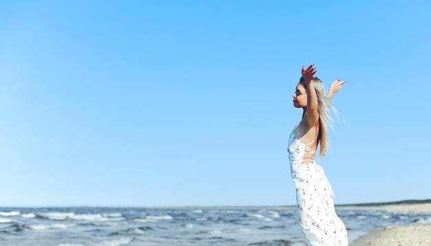 写真 白いサマードレスを着て腕を広げ、海のビーチに立っている幸せな金髪の美しい女性