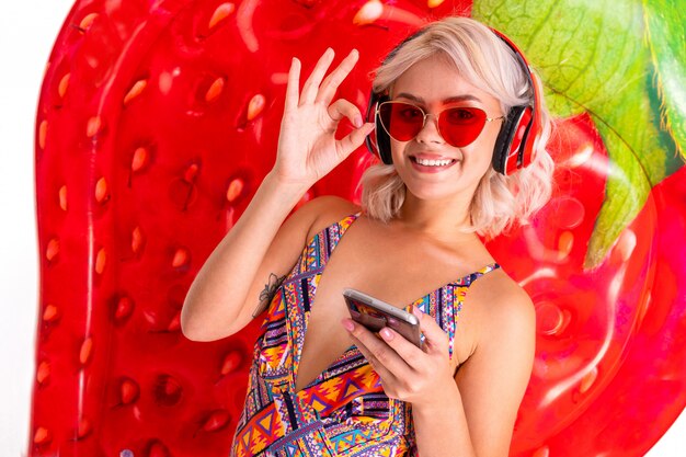 Happy blonde caucasian female stands in swimsuit with big rubber mattress, phone, earphones and smiles isolated