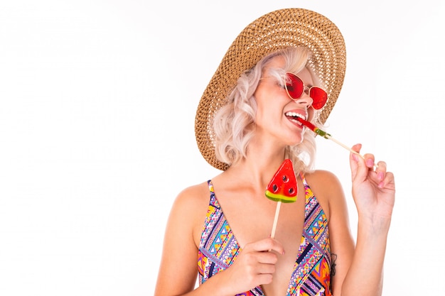 Happy blonde caucasian female stands in swimsuit with watermelon lolipops and smiles isolated