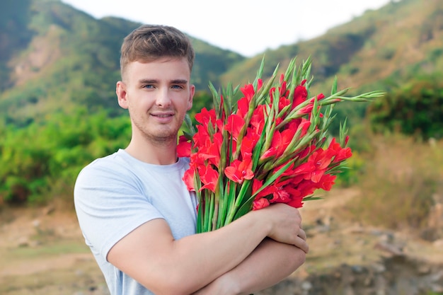 幸せな金髪ヨーロッパのハンサムな男、赤いscar色の花の美しい花束を持つロマンチックな若者、グラジオラスまたはグラジオラスアウトドア、笑みを浮かべて。デート、プレゼント、ギフト、花のコンセプト。
