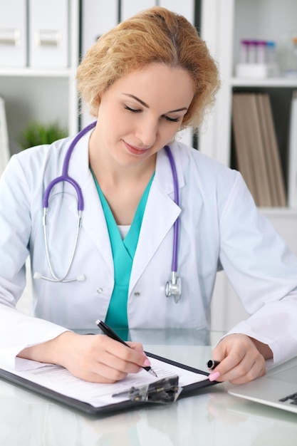 Happy blonde female doctor sitting at the table and filling up medical history form Medicine healthcare and help concept Physician ready to examine patient