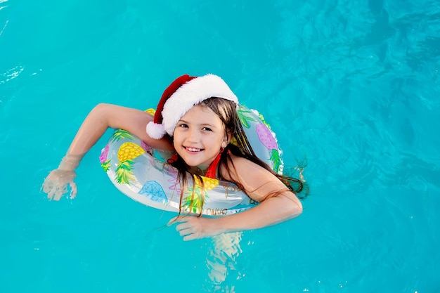 a happy blonde girl in a Santa Claus hat and a lifebuoy is swimming in the pool