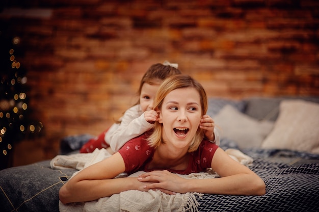 Happy blonde hair mother in red pajama with joking daughter on her back lying on grey bed looking sideways in front of brown brick wall and christmas tree with lights of garland