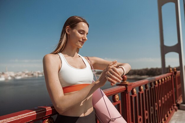 Happy blonde lady with mat checks time on smartwatch on contemporary footbridge