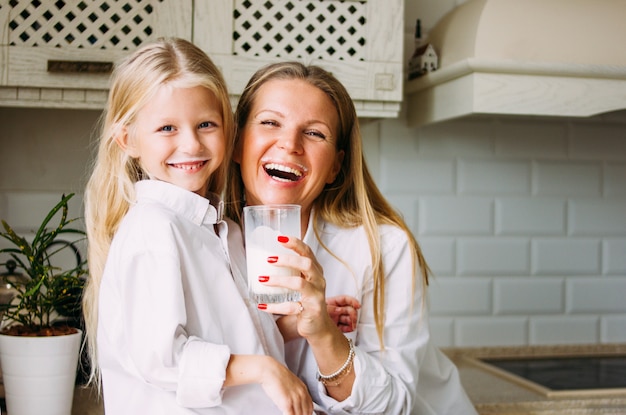 幸せなブロンドの長い髪のママと娘の明るいキッチン、健康的なライフスタイルで牛乳を飲む
