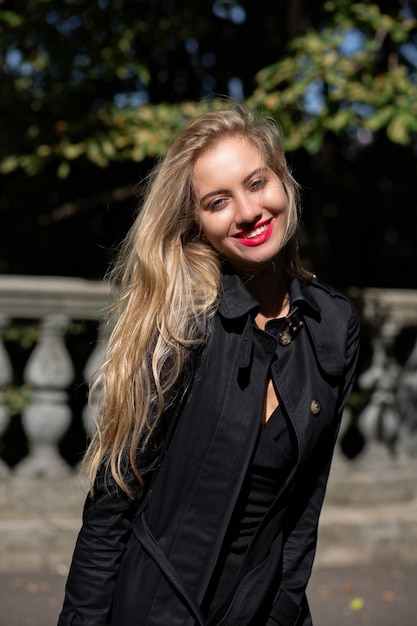 Happy blonde model wearing black coat and dress posing in rays of sun at the city