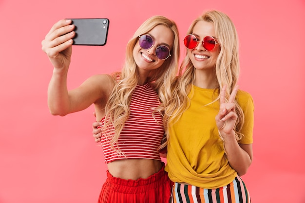 Happy blonde twins in sunglasses having rest and making selfie on smartphone over pink wall