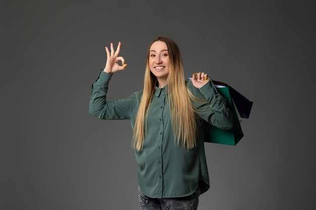 Happy blonde woman holding shopping bags with purchases and showing Ok gesture on gray background.