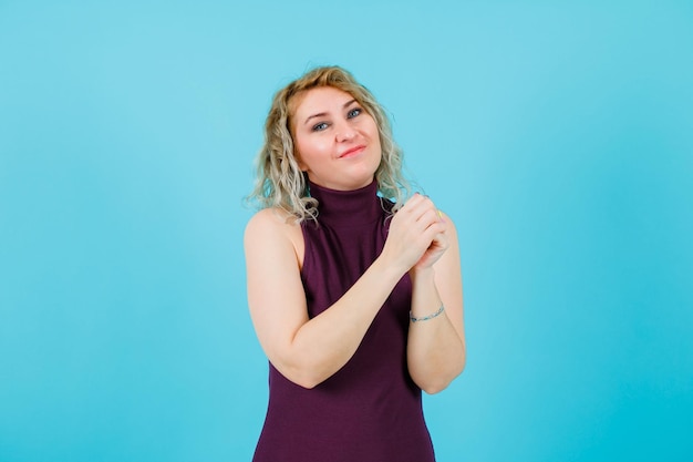 Happy blonde woman is looking at camera by holding hands together on blue background