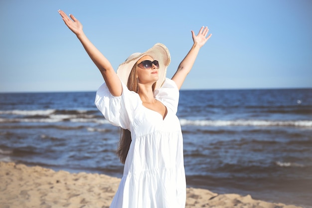 Happy blonde woman is on the ocean beach in a white dress, sunglasses and hat, raising hands.