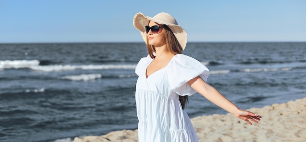 행복한 금발의 여인은 흰 드레스와 선글라스를 끼고 팔을 벌리고 바다 해변에 있습니다.