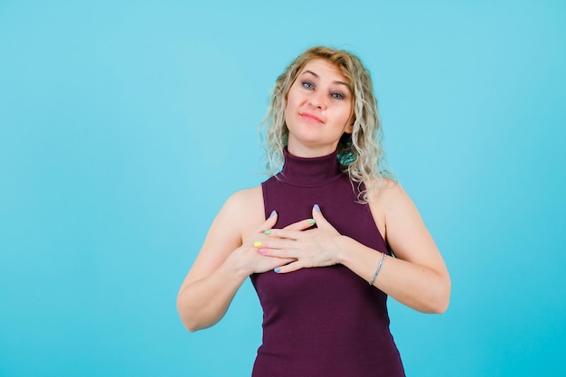 Foto la donna bionda felice sta posando alla macchina fotografica tenendo le mani insieme sul petto su fondo blu