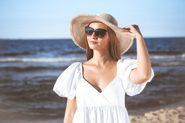 행복한 금발 여성이 선글라스와 모자를 들고 바다 해변에서 포즈를 취하고 있습니다. 저녁 태양