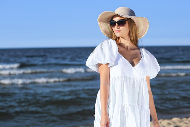 Happy blonde woman is posing on the ocean beach with sunglasses and a hat. Evening sun.