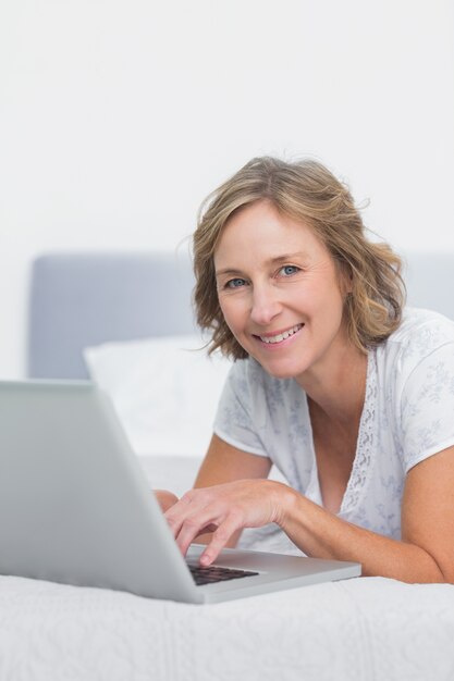 Happy blonde woman lying on bed using laptop