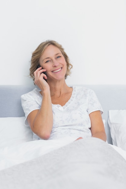 Счастливый блондинка женщина по телефону в постели, улыбка на камеру