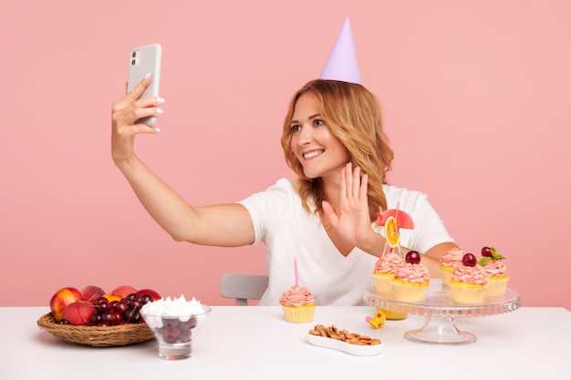 Felice donna bionda che registra video per il suo vlog di cibo utilizzando lo smartphone per trasmettere in livestream agitando la mano alla telecamera e sorridendo in studio indoor girato isolato su sfondo rosa