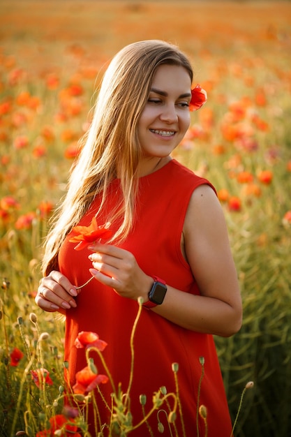 Happy blonde woman in red dress holding a flower in her hair on a poppy field. Fun summer vibes. Relaxed atmosphere, positive emotions.