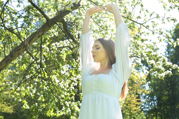 Счастливая блондинка в белом платье на фоне цветущего весеннего сада с поднятыми к небу руками