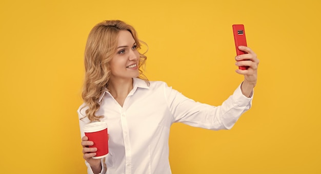 黄色の背景に電話でselfieを作るコーヒーカップと幸せなブロンドの女性