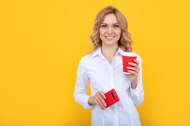 Felice donna bionda con tazza di caffè e smartphone su sfondo giallo