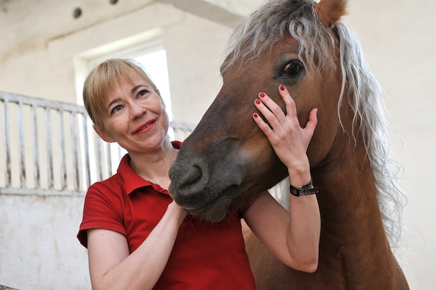 Happy blonde woman with a horse in a stable on the ranch.
