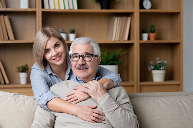 Happy blonde young woman embracing her cheerful senior father on couch while both looking at you during home relax