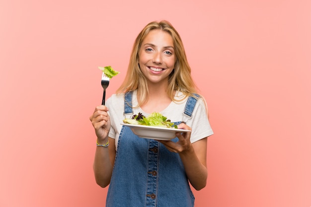 Foto giovane donna bionda felice con insalata sopra la parete rosa isolata