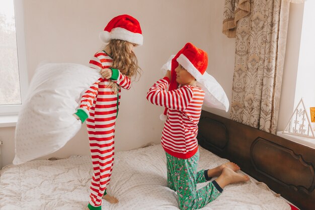 クリスマスの朝にクリスマスの帽子をかぶった幸せな男の子と女の子がベッドで枕と遊んで戦う