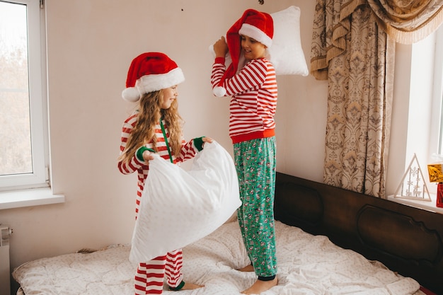 Happy boy and girl in christmas hats on christmas morning play and fight with pillows in bed