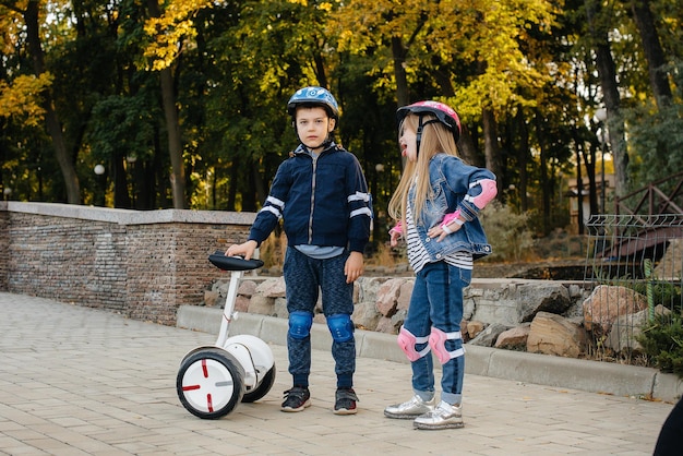 幸せな男の子と女の子が立って、日没時にセグウェイとスクーターの公園に乗ります。幸せな子供時代。