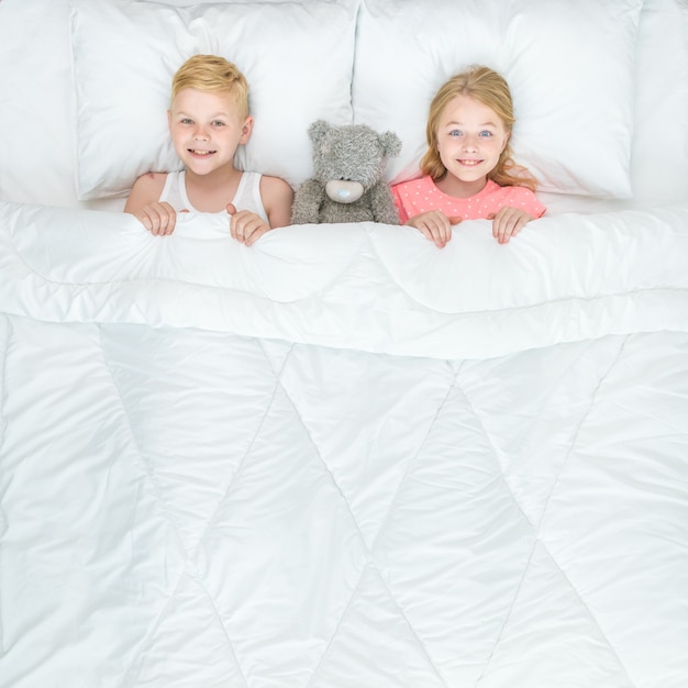 The happy boy and girl with a soft toy lay on the bed. view from above