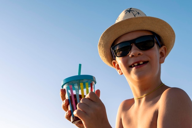 Happy boy in hat and sunglasses drinking juice on the beach