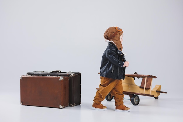 Счастливый мальчик в шлеме и куртке пилота несет коричневый чемодан Портрет ребенка-пилота Ребенок в кожаной куртке Маленький путешественник с чемоданом