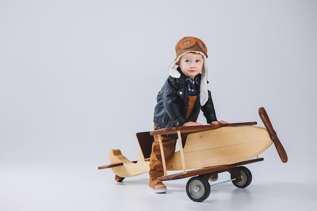 A happy boy in a helmet and a pilot\'s jacket stands near a\
wooden plane portrait of a child pilot a child in a leather jacket\
wooden toys eco plane from tree