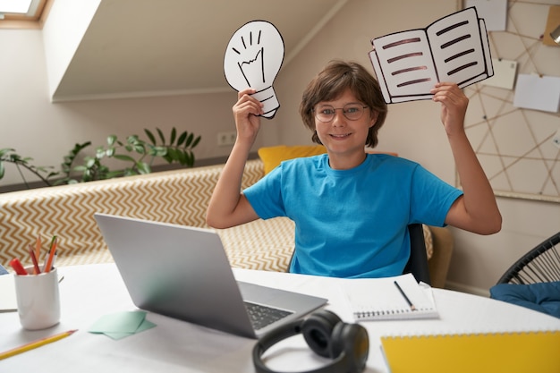 Happy boy holding paper light bulb and book looking at camera and smiling while learning online at