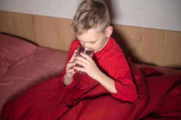 A happy boy is sitting in a red bed under a blanket and drinking water from a glass