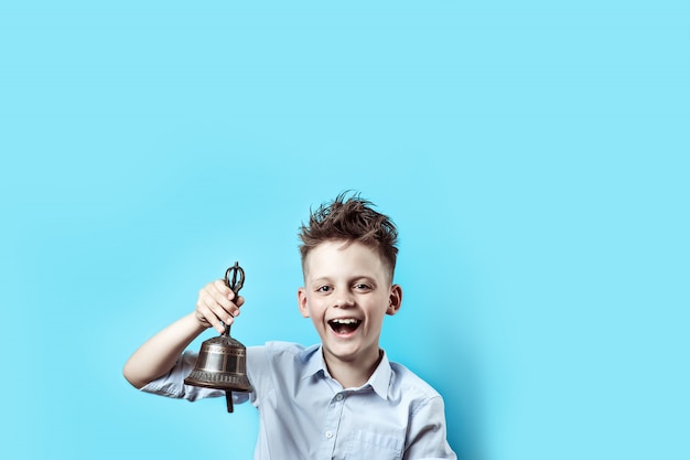 A happy boy in light shirt goes to school. he has a bell in his hand, which he rings and smiles.