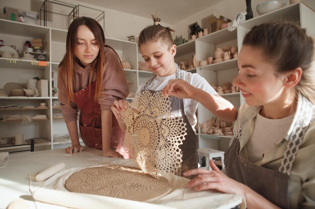 Happy boy making a pattern on a piece of clay during pottery class with professional ceramist and his mom